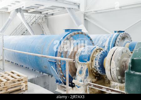 Partie d'une énorme machine industrielle avec un grand arbre métallique bleu pendant le processus de production de marbre dans un atelier ou une usine spacieux et blanc Banque D'Images