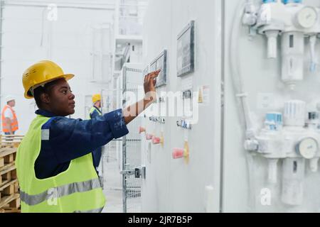 Jeune Afro-américaine en uniforme et hardhat en appuyant sur le bouton sur le panneau de commande tout en allant démarrer la machine de production Banque D'Images