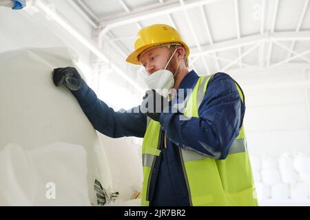 Ingénieur de l'usine moderne ou de l'usine de production mettant le respirateur sur son visage tout en se tenant devant un énorme sac blanc avec des matières premières Banque D'Images