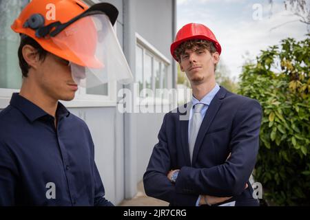 Portrait d'un ingénieur au look assuré et bras pliés portant un casque de sécurité pendant que son collègue travaille Banque D'Images
