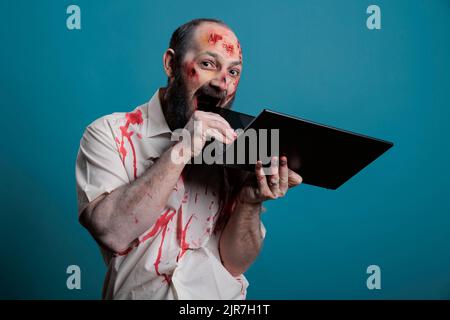 Un zombie effrayant piquant sur un ordinateur portable, ayant des cicatrices sanglantes et des blessures sur un visage sale et effrayant. Effrayant monstre mort-undead étant agressif avec pc, l'aspect dangereux cruel et mortel. Banque D'Images