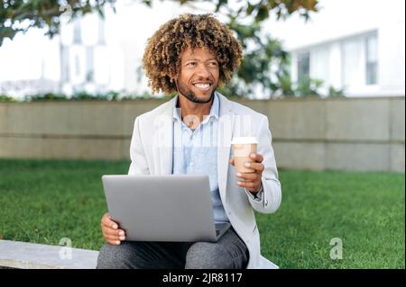 Bon positif brésilien ou espagnol réussi, employé de l'entreprise, entrepreneur, assis à l'extérieur avec un ordinateur portable dans ses mains, tient un verre de café à la main, pauses au travail, regarde loin, sourit Banque D'Images