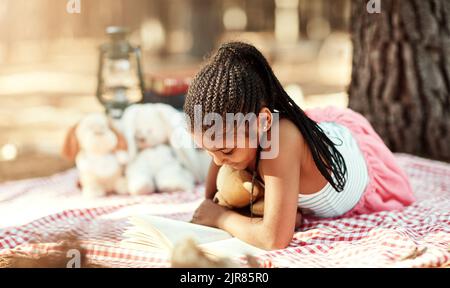 Son endroit préféré pour apprécier une bonne histoire. Une petite fille lisant un livre avec ses jouets dans les bois. Banque D'Images