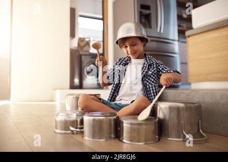 HES un peu bruyant. Un petit garçon heureux jouant des tambours avec des pots sur le sol de la cuisine tout en portant un bol sur sa tête. Banque D'Images