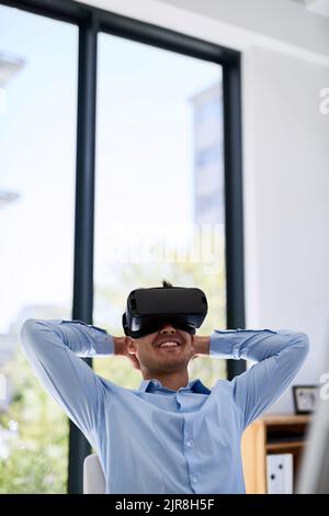 Il peut désormais réfléchir à une vue sur la plage. Un jeune homme d'affaires heureux portant un casque de réalité virtuelle tout en travaillant à son bureau. Banque D'Images