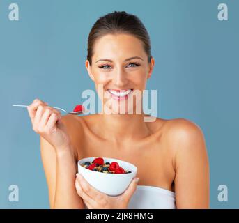 "Quand je mange bien, je me sens bien." Studio photo d'une jeune femme attrayante mangeant un bol de muesli et de fruits sur fond bleu. Banque D'Images