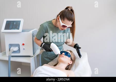 Thérapeute le cosmétologue subit un traitement au laser sur le visage jeune femme en clinique de beauté. Poils laser du visage. Procédures d'épilation. Client satisfait, attention sélective. Banque D'Images