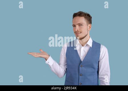 Jeune homme surpris montrant le geste de la main en copie vierge. Surpris homme debout à côté regardant l'appareil photo portant une chemise blanche et veste bleue isolée sur fond bleu Banque D'Images