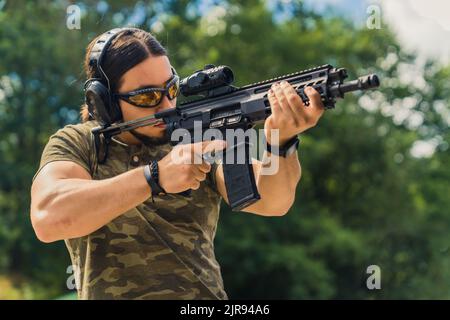 Beau moniteur masculin à l'aire de tir. Tir de plein air moyen d'un homme musclé orienté caucasien dans des lunettes de protection et des écouteurs visant la cible avec un fusil regardant à travers la loupe. Photo de haute qualité Banque D'Images