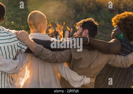 Vue arrière de multiculturel bon vieux amis qui embrasse et d'avoir un pique-nique de fin de semaine dans la nature près du feu de camp, parler et rire. Heure du coucher du soleil. Photo de haute qualité Banque D'Images