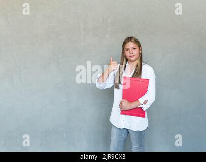 Une jeune femme suit des cours, une étudiante étudie, tient des carnets et montre un pouce vers le haut dans l'approbation, recommander. Photo de haute qualité Banque D'Images