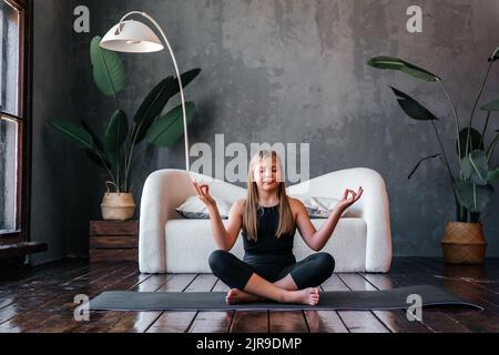 Jeune fille attrayante pratiquant le yoga, assis dans la moitié de l'exercice de Lotus, Ardha Padmasana pose, exercice, session de méditation, intérieur de maison. Banque D'Images