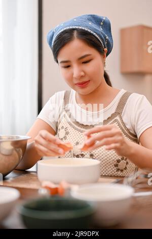 Belle jeune asiatique femelle crack un oeuf et séparer le jaune d'oeuf de blanc d'oeuf, en préparant les ingrédients du cupcake dans la cuisine. Cupcake recette conc Banque D'Images