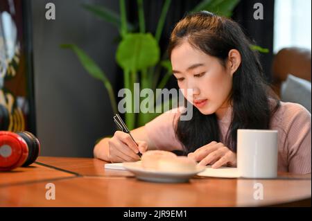 Une charmante jeune étudiante asiatique se trouve dans le café, se concentrant sur ses devoirs, écrivant quelque chose sur son carnet. Banque D'Images