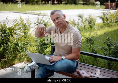 Homme âgé assis sur un banc en bois avec une jambe croisée dans le parc en été, tenant un ordinateur portable ouvert, pointant l'index vers le bas. Banque D'Images