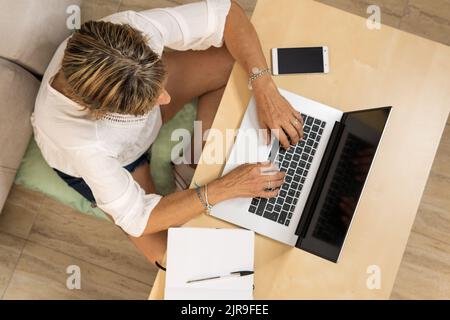 Femme mature travaillant à la maison dactylographiant sur ordinateur portable avec agenda et téléphone portable sur table en bois. Vue aérienne Banque D'Images