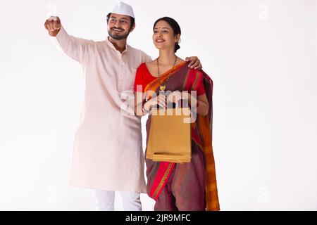 Portrait d'un couple Maharashtrian debout avec des sacs de shopping et pointant vers l'extérieur Banque D'Images