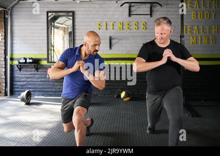 Entraîneur personnel masculin instruisant et aidant l'homme mature senior tout en faisant avant fente de jambe exercice dynamique de mouvement dans la salle de gym à la maison petite entreprise Banque D'Images