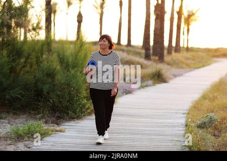 Femme active d'âge moyen avec tapis de yoga et marchant à la plage pour pratiquer le yoga, s'exerçant à l'extérieur avec le sentiment de bonheur au lever du soleil le matin. Co Banque D'Images