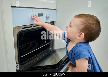 Bébé monte dans un four électrique chaud. Un enfant ouvre la porte du four dans la cuisine. Enfant d'un an Banque D'Images