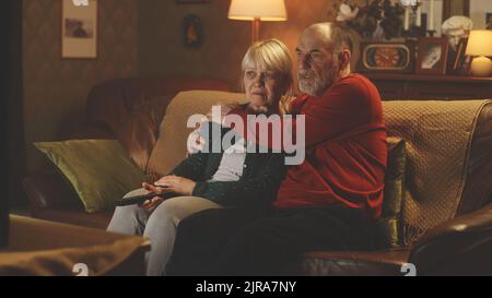 Homme âgé embrassant une femme pleurant tout en se reposant sur un canapé et en regardant un film triste à la télévision ou les nouvelles du soir, le soir dans un salon confortable à la maison Banque D'Images