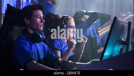 Jeune femme gaie, joueur esportsman dans des lunettes et des écouteurs souriant et donnant des cinq hauts, à des coéquipiers masculins tout en célébrant la victoire dans le match de jeu vidéo pendant le tournoi professionnel Banque D'Images