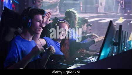 Jeune femme gaie, joueur esportsman dans des lunettes et des écouteurs souriant et donnant des cinq hauts, à des coéquipiers masculins tout en célébrant la victoire dans le match de jeu vidéo pendant le tournoi professionnel Banque D'Images