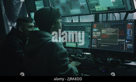 Deux hackers dans des vêtements décontractés craquant des fichiers classifiés sur l'ordinateur et infectant des documents avec un virus dans une pièce sombre avec des moniteurs Banque D'Images