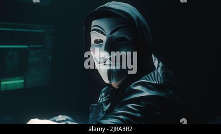Homme dans un masque anonyme exécutant une cyber-attaque de pirate sur les serveurs d'entreprise et l'ordinateur et regardant la caméra tout en étant assis au bureau dans l'obscurité Banque D'Images