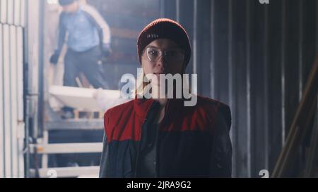 Femme travailleuse dans la veste et le chapeau regardant la caméra contre l'entrée de l'atelier tandis que les collègues déchargent le camion avec des matériaux Banque D'Images