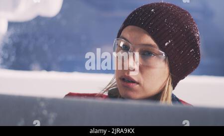 Employée féminine dans un chapeau et des lunettes qui retire la poussière de plastique du bloc polymère pendant le travail dans un atelier professionnel moderne Banque D'Images