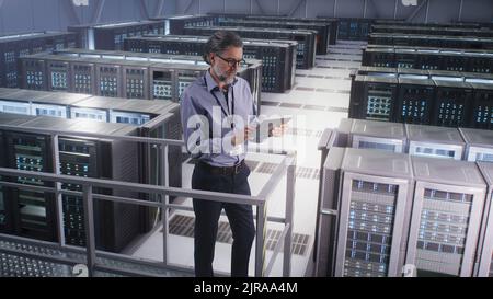 Un spécialiste informatique utilise Digital Pad dans un centre de Cloud Computing ou un centre minier de Big Server Farm, pour surveiller la cybersécurité et l'état des serveurs. Panoramique grand angle autour de la vue. Banque D'Images