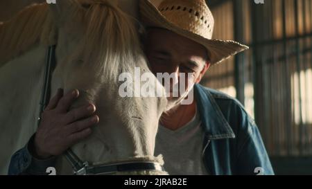 Homme senior positif en chapeau de paille embrassant et embrassant la tête du cheval albino tout en travaillant en stable sur le ranch Banque D'Images