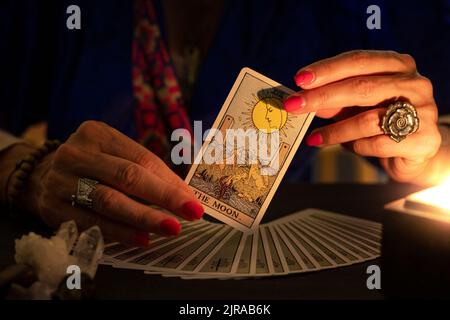 Les mains de la caissière montrant la carte de la Lune tarot, symbole de l'intuition, pendant une lecture. Gros plan avec bougie, ambiance feutrée. Banque D'Images