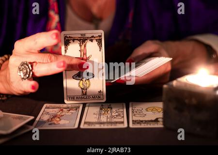 Les mains de femmes de la caissière montrant la carte de tarot d'homme pendu, symbole de l'attente, pendant une lecture. Gros plan avec bougie, ambiance feutrée. Banque D'Images