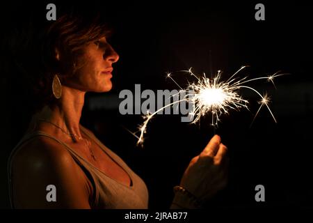 Femme caucasienne attirante tenant un sparkler brûlant ou bengale dans l'obscurité. Portrait de profil. Vacances ou fond magique ou papier peint. Banque D'Images