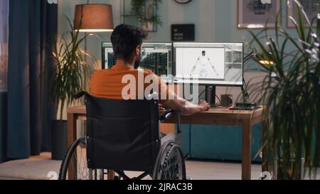Vue arrière d'un développeur masculin méconnaissable avec un handicap assis sur un fauteuil roulant et rendant le modèle 3D de robot sur un ordinateur pendant le travail dans un salon sombre à la maison Banque D'Images