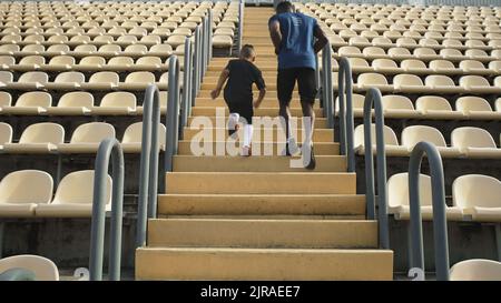 Vue arrière d'un père et d'un fils méconnaissables qui s'exécutent sur un escalier près des sièges pendant l'entraînement, le jour ensoleillé du stade Banque D'Images