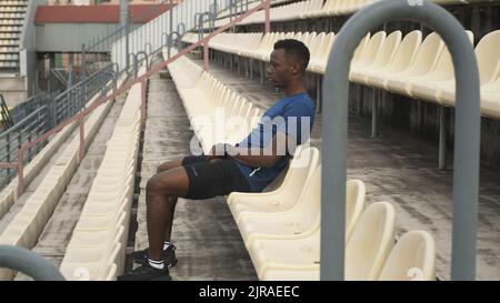 Zoomez sur la vue d'un sportif noir fatigué assis sur un siège en plastique et de se détendre pendant une pause dans un entraînement au stade Banque D'Images