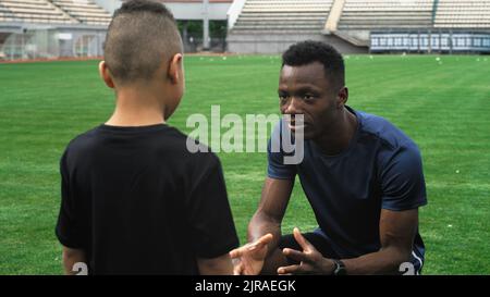 Zoom arrière de l'homme noir parlant avec un garçon de course mixte pratiquant avec le ballon pendant l'entraînement de football sur le stade Banque D'Images