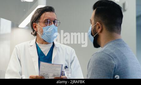 Médecin de sexe masculin d'âge moyen en masque parlant et donnant une ordonnance à l'homme tout en travaillant dans une clinique moderne pendant une pandémie Banque D'Images
