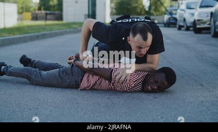 Femme agressive en uniforme de police appuyant sur l'homme noir au sol et parlant pendant l'arrestation dans la rue Banque D'Images