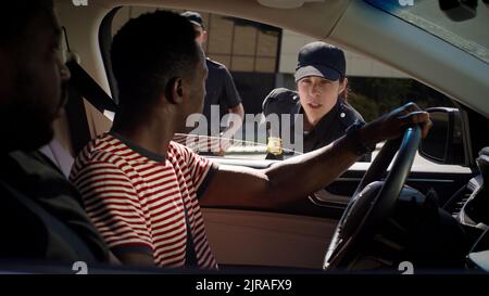 Des hommes afro-américains se gesticulent et discutent de la présence d'une femme sérieuse en uniforme de police lorsqu'elle est assise en voiture après vérification des documents Banque D'Images
