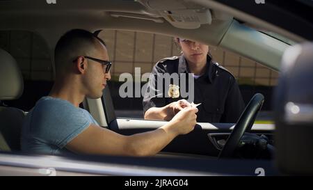 Femme sérieuse dans le permis de contrôle d'uniforme de police et de poser des questions à l'homme assis dans la voiture sur la rue de ville Banque D'Images