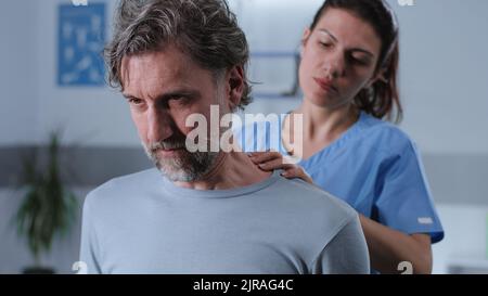 Femme adulte en uniforme médical massant le cou et les épaules d'un homme d'âge moyen barbu tout en travaillant dans une clinique de réadaptation Banque D'Images