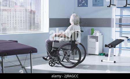 Un homme anonyme âgé ayant une déficience assis sur un fauteuil roulant et regardant la fenêtre après une séance de réadaptation à l'hôpital Banque D'Images