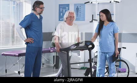 Les médecins masculins et féminins aidant l'homme âgé à grimper sur un tapis roulant et expliquant l'exercice de réadaptation pendant le travail dans la clinique moderne Banque D'Images