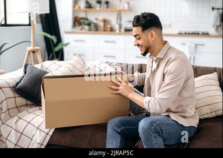 Excitée course mixte jeune homme ouvrant grande boîte en carton, déballant la commande de magasin d'Internet à la maison, heureux avec le service de livraison rapide et les marchandises qui ont reçu, sourire, heureux des émotions faciales Banque D'Images