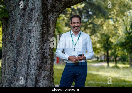 Homme d'affaires heureux tenant un smartphone debout près d'un arbre Banque D'Images