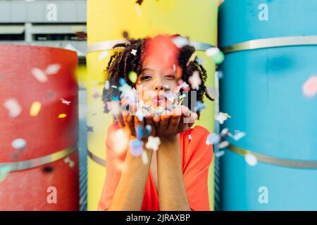 Jeune femme souriante soufflant des confettis devant des tuyaux colorés Banque D'Images